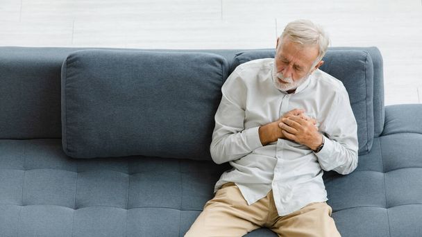 Kaukázusi idős, idős, ősz haj és szakállas férfi, aki szorosan a kanapén fekszik a nappaliban, egyedül, kéz a kézben a mellkason, hirtelen problémától szenvedve, fájdalmas szívroham.. - Fotó, kép