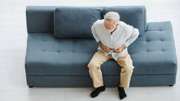 Caucasian old senior elderly stressed unhealthy gray hair and bearded man sitting on sofa in living room at home alone holding hands on belly suffering from painful indigestion diarrhea stomachache. - Photo, Image