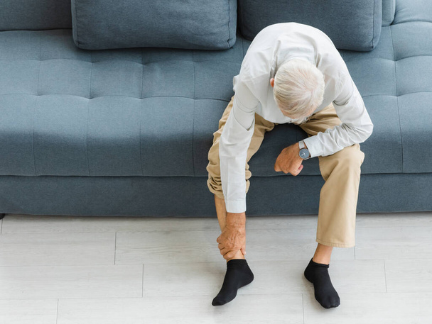 Kaukázusi idős idős, egészségtelen ősz hajú férfi ül kanapén a nappaliban otthon egyedül hajlító lefelé mutató kéz a bokán szenved fájdalmas sérülés feszültség ínszalag és ízületi fájdalom. - Fotó, kép