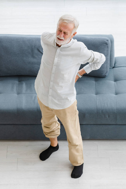Kaukázusi idős idős idős, egészségtelen, beteg ősz haj és szakállas férfi áll közel kanapé a nappaliban otthon egyedül fogja a kezét a hátán szenved fájdalmas sérülés törzs fáj gerinc hátfájás. - Fotó, kép