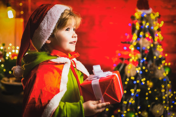 Happy little boy by near Christmas tree with Christmas gift. Little kid is wearing Santa clothes. Christmas kids concept. - Фото, изображение