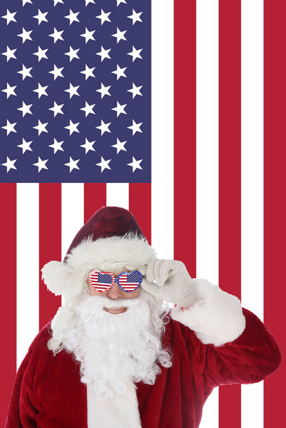 Le Père Noël sourit en enfilant ses lunettes de soleil avec un fond drapeau américain. Joyeux Noël et Joyeuses Fêtes. Joyeux Noël et bonne année. Carte de voeux avec le Père Noël assis devant le drapeau américain. Vacances d'hiver. Ho Ho Ho. - Photo, image
