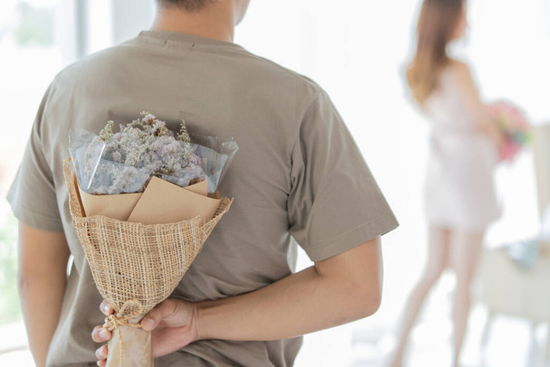 A man wearing brown t shirt holding and hiding white flowers bouquet behind his back when walking to his long hair girlfriend wearing sleeveless dress in blurred background. - Foto, afbeelding