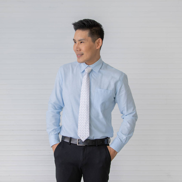 Portraitaufnahme eines asiatischen männlichen Models mit kurzen schwarzen Haaren, das ein hellblaues Hemd mit weißer Punktekrawatte trägt, lächelnd vor weißem Streifenhintergrund seine Hände in eine schwarze Hosentasche legt. - Foto, Bild