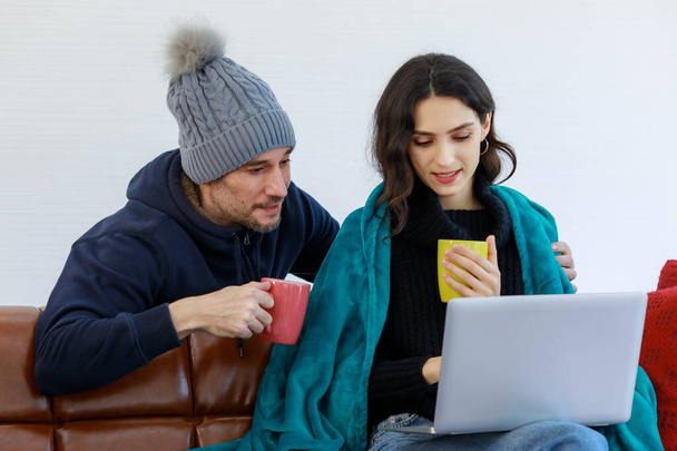 Caucasian young bearded husband in jacket knitted hat and pretty wife cover by blanket sit on couch drink hot coffee when browsing surfing internet from laptop computer together in winter at home. - Photo, Image
