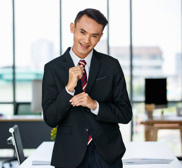 Позитивний азіатський чоловік-підприємець коригує костюм, стоячи в сучасному робочому місці і дивлячись на камеру - Фото, зображення