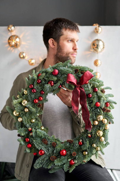 Christmas wreath of fresh spruce in man hands. Xmas circlet with red and gold ornaments and balls. Christmas mood. Gray wall on background. - Photo, Image