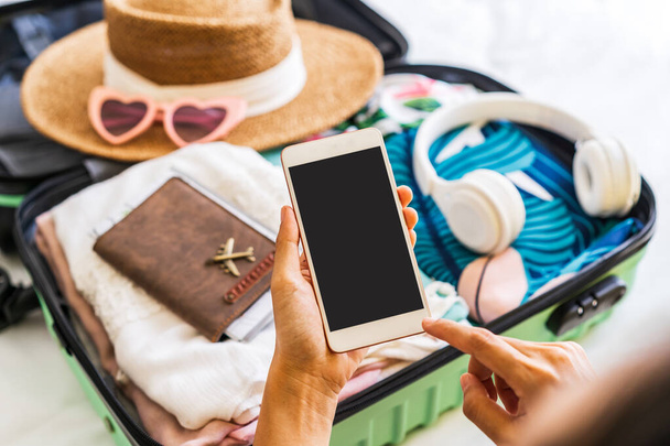 Young woman traveler sitting on the bed using smartphone and packing her suitcase preparing for travel on summer vacation - Foto, Imagen