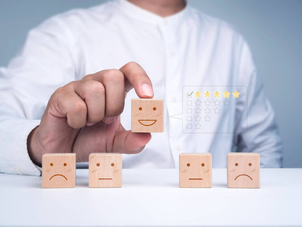 Customer service evaluation, ratings, feedback, client experience, and satisfaction survey concepts. The man in a white shirt picks the happy face wooden block with a check mark on five stars rating. - Photo, Image