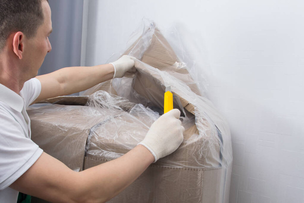 the delivery man in protective gloves, opens the furniture from the box, cuts the dense transparent polyethylene, with a stationery knife - Fotoğraf, Görsel
