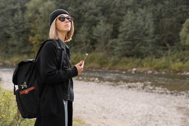 Caucasian girl tourist traveling near the river with a backpack, nature background, stylish young woman traveler in a black coat, side view. Tourism and travel concept. - Photo, Image