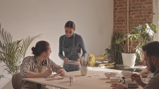 Langzaam shot van kleine groep mensen zitten rond tafel tijdens keramiek masterclass in gezellige kunststudio. Vrouwelijke pottenbakker legt uit hoe keramische artikelen te schilderen - Video