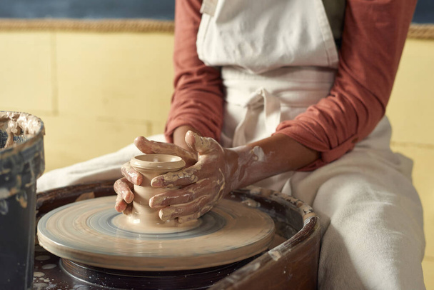 Colegiala en delantal sosteniendo olla de arcilla en la rueda de cerámica giratoria - Foto, Imagen