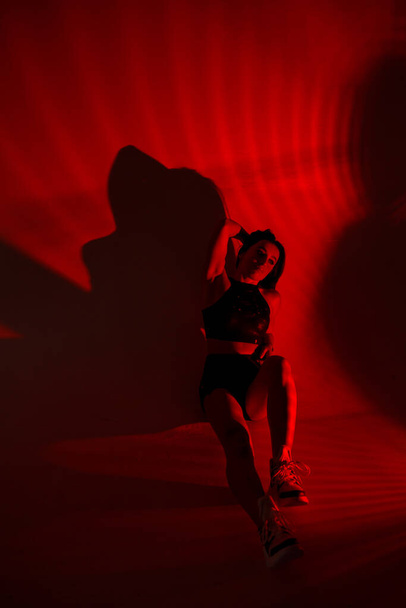 Photoshoot of a young brunette in a photo studio. Beautiful slender woman in a black top and shorts in red light. - Φωτογραφία, εικόνα