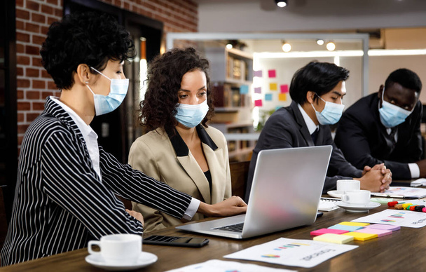 Wielorasowe mieszane grupy biznesowe pracujące z koncentracją w biurze, noszące maski twarzy jako nowe normalne, aby chronić lub zapobiegać wirusom w czasie pandemii, używając laptopa i dyskusji o pracy. - Zdjęcie, obraz