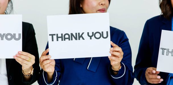 Studio shot of stripe letters thank you paper sign holding by smiling female officer in business suit at chest showing appreciation with faceless colleagues to customers on white background. - Foto, imagen