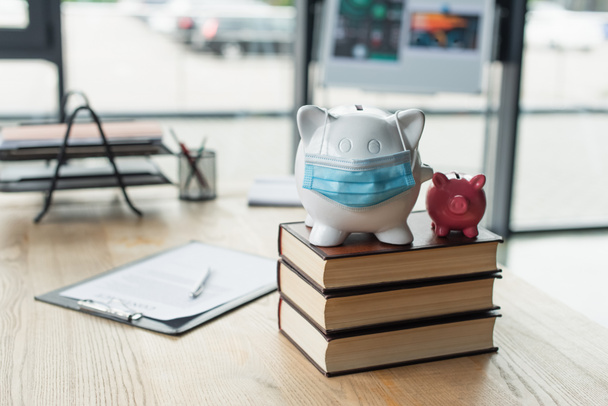 piggy banks with medical mask on codex books near blurred contract on desk, anti-corruption concept - Photo, Image