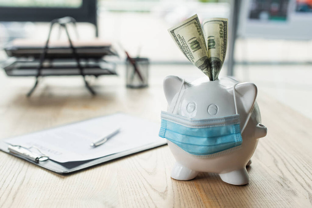 medical mask on piggy bank with dollar banknotes near blurred contract on desk, anti-corruption concept - 写真・画像