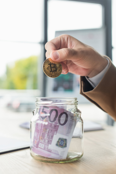 KYIV, UKRAINE - SEPTEMBER 22, 2021: cropped view of businessman holding bitcoin near glass jar with euro banknotes, anti-corruption concept - 写真・画像
