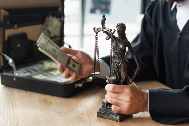 vista parziale di avvocato in possesso di statuetta e dollari vicino valigetta sfocata, concetto di anticorruzione - Foto, immagini
