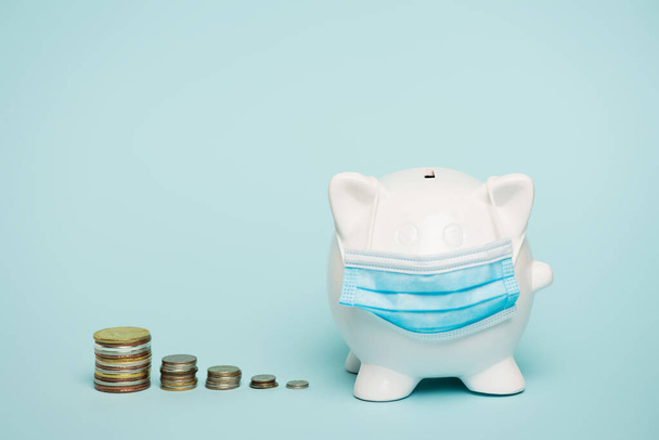stacks of coins near piggy bank in medical mask on blue background with copy space, anti-corruption concept - Photo, Image