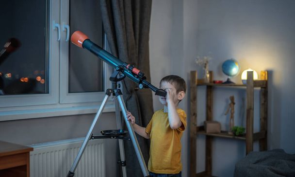 Cute boy is looking through a telescope in a room at the night starry sky. Children's scientific hobbies and space exploration - Photo, image