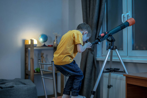 Cute boy is looking through a telescope in a room at the night starry sky. Children's scientific hobbies and space exploration - Photo, image
