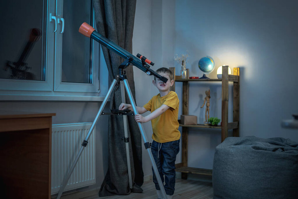 Cute boy is looking through a telescope in a room at the night starry sky. Children's scientific hobbies and space exploration - Fotoğraf, Görsel