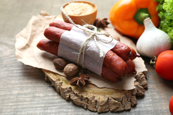Composition of smoked thin sausages, mustard in bowl and spices on cutting board, on wooden background - Фото, зображення