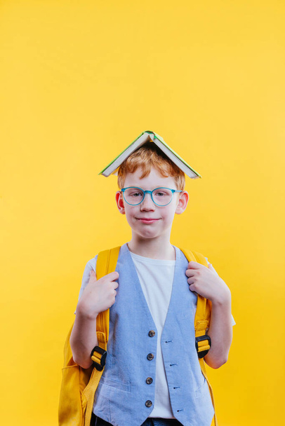 Αστεία κόκκινα μαλλιά σχολιαρόπαιδο με γυαλιά μάτια κρατώντας βιβλίο στο κεφάλι του ως στέγη σε κίτρινο φόντο με κενό χώρο για κείμενο - Φωτογραφία, εικόνα