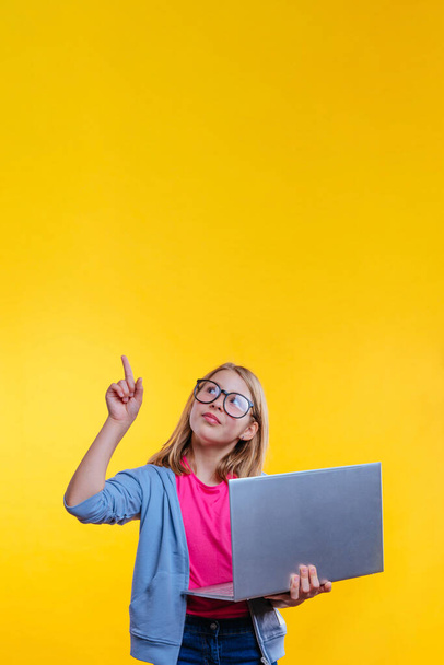 Szczęśliwa blondynka włosy uczennica z okularami trzyma laptopa i wskazując palcem na żółtym tle z pustym miejscem na tekst - Zdjęcie, obraz