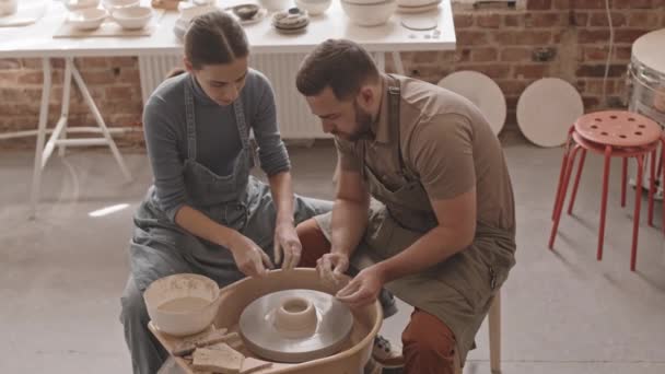 若い女性陶芸家のパンニングメディアのクローズアップ居心地の良いワークショップで陶器の車輪で一緒に座って土鍋を作るために男を教えて - 映像、動画