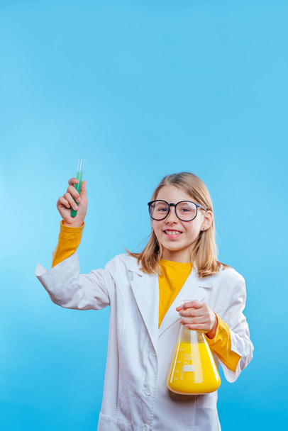 Školačka drží chemickou nádobu a zkumavky na sobě ochranné šaty a brýle na modrém pozadí s prázdným prostorem pro text - Fotografie, Obrázek