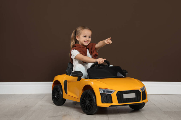 Cute little girl driving children's electric toy car near brown wall indoors - Foto, Bild