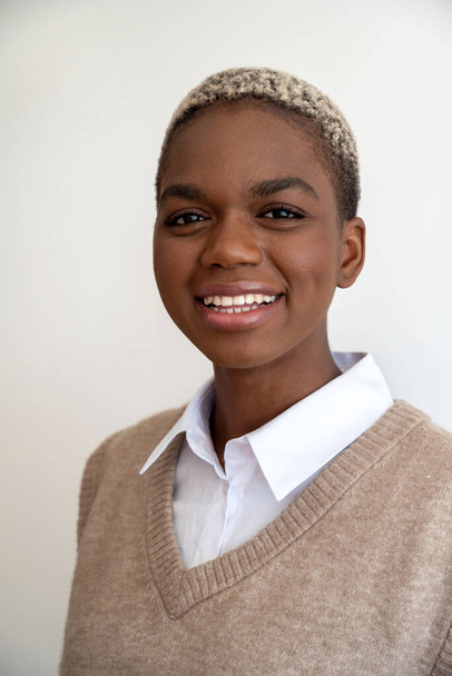Pozitivní Afroameričan ve stylovém oblečení a s barvenými krátkými vlasy při pohledu na kameru s okouzlujícím úsměvem na bílém pozadí ve studiu - Fotografie, Obrázek