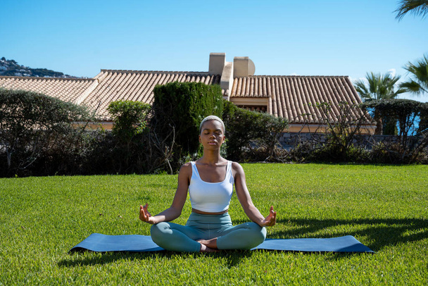 Entspannte Afroamerikanerin in Aktivkleidung sitzt auf einer Matte auf einer Wiese in Padmasana und meditiert mit geschlossenen Augen und Mudra-Händen während einer Yoga-Stunde im Sommerpark - Foto, Bild