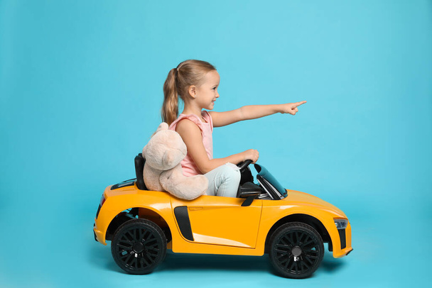 Cute little girl with toy bear driving children's car on light blue background - Photo, image