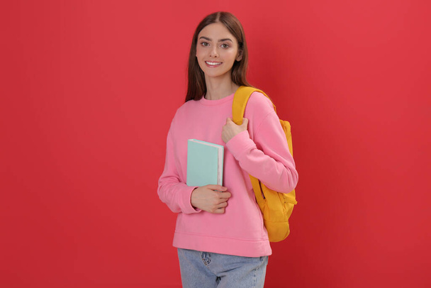 Teenage student with backpack and book on red background - Фото, зображення