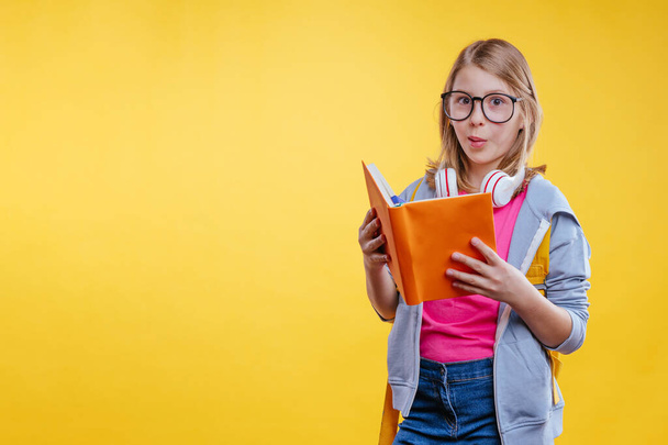 Happy teenager girl holding backpack reading book on yellow background - Foto, afbeelding