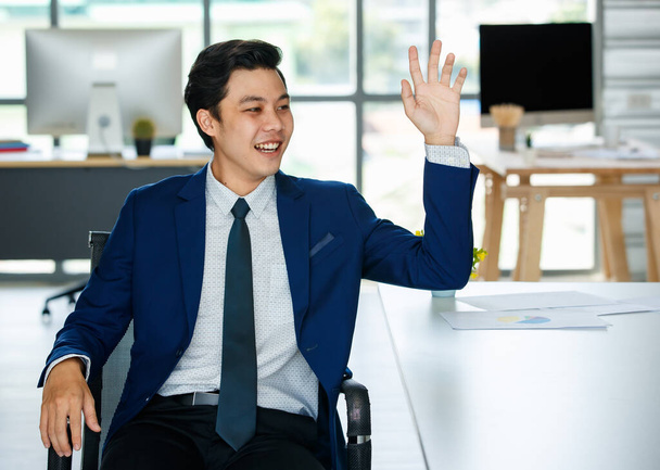 Fiatal és elegáns ázsiai üzletember kék öltönyben ül a modern irodában és felemelt kézzel üdvözöl vagy köszön valakinek.. - Fotó, kép