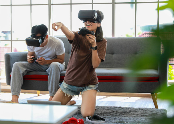 Joven pareja asiática atractiva sosteniendo joysticks disfrutar jugando videojuegos juntos con gafas VR con diversión y emoción en la sala de estar con ventanas de vidrio de fondo. - Foto, imagen