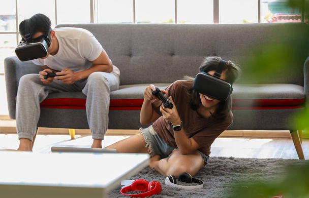 Νεαρό ελκυστικό ασιατικό ζευγάρι κρατώντας joysticks απολαμβάνουν να παίζουν βιντεοπαιχνίδια μαζί φορώντας γυαλιά VR με διασκέδαση και ενθουσιασμό στο σαλόνι με τα παράθυρα από γυαλί φόντο. - Φωτογραφία, εικόνα
