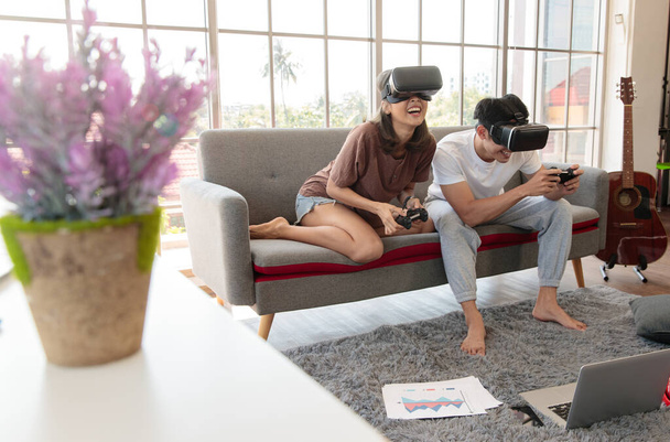 Joven pareja asiática atractiva sosteniendo joysticks disfrutar de saltar y jugar a videojuegos juntos con gafas VR con diversión y emoción en la sala de estar con ventanas de vidrio de fondo. - Foto, imagen