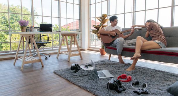 Joven pareja asiática atractiva sentados juntos en un sofá. Hombre tocando la guitarra y la mujer escuchando y sonriendo en la sala de estar con ventanas de vidrio de fondo. Concepto de amor y relación de pareja. - Foto, imagen