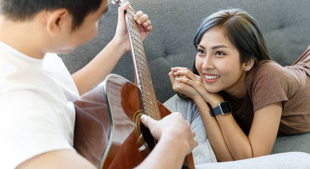 Young attractive Asian couple together on a couch. Man sitting playing guitar and woman lying and listening in living room with glasses window background. Concept for love and couple relationship. - Foto, afbeelding