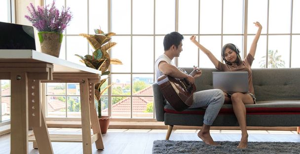 Giovane attraente coppia asiatica seduta insieme su un divano. Un uomo che suona la chitarra e una donna che guarda un portatile con le cuffie. Concetto di amore e relazione di coppia. - Foto, immagini