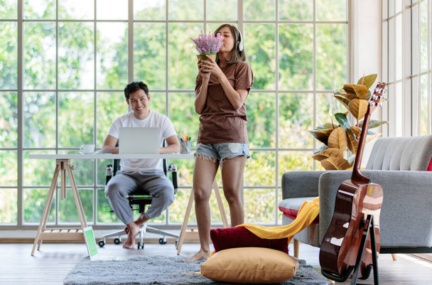 Fiatal vonzó ázsiai pár együtt élnek a nappaliban. Nő kezében és illata vödör virág és férfi ül az asztalon dolgozik laptop. A szerelem és a párkapcsolat fogalma. - Fotó, kép