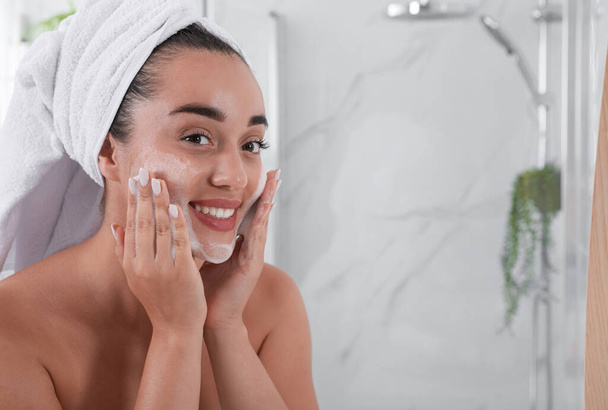 Beautiful young woman applying cleansing foam onto face in bathroom, space for text. Skin care cosmetic - Foto, Imagen
