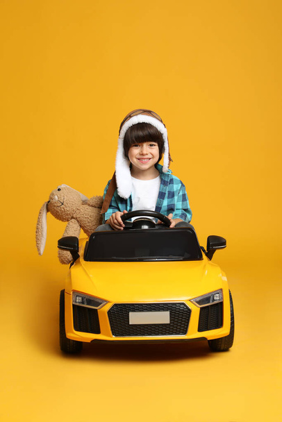 Cute little boy with toy bunny driving children's car on yellow background - Foto, afbeelding