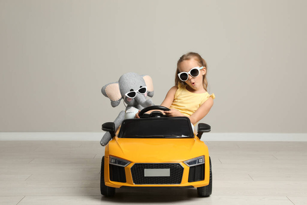 Cute little girl with toy elephant driving children's car near grey wall indoors - Φωτογραφία, εικόνα